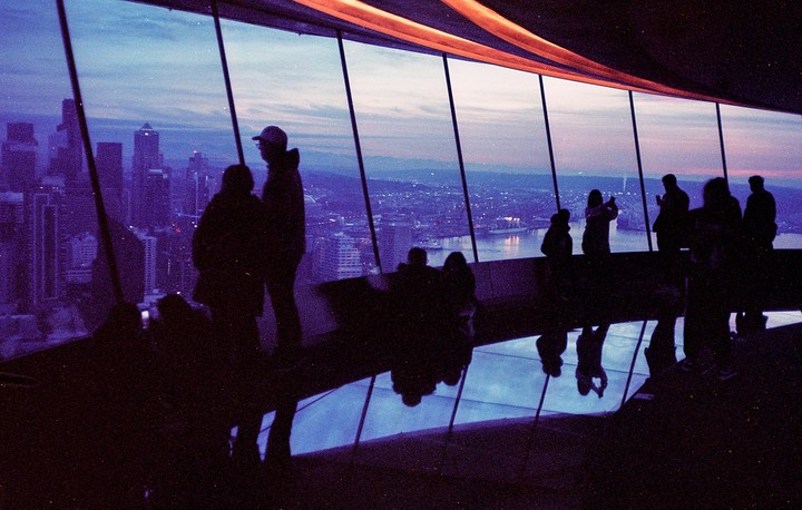 Silhouetted people look out from a tall building at a city and water at dusk.