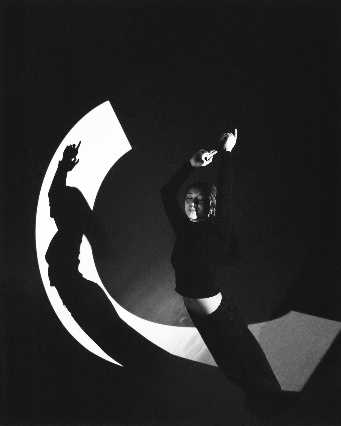 A woman stands in a dark room with a crescent-shaped light and her shadow cast on the wall.