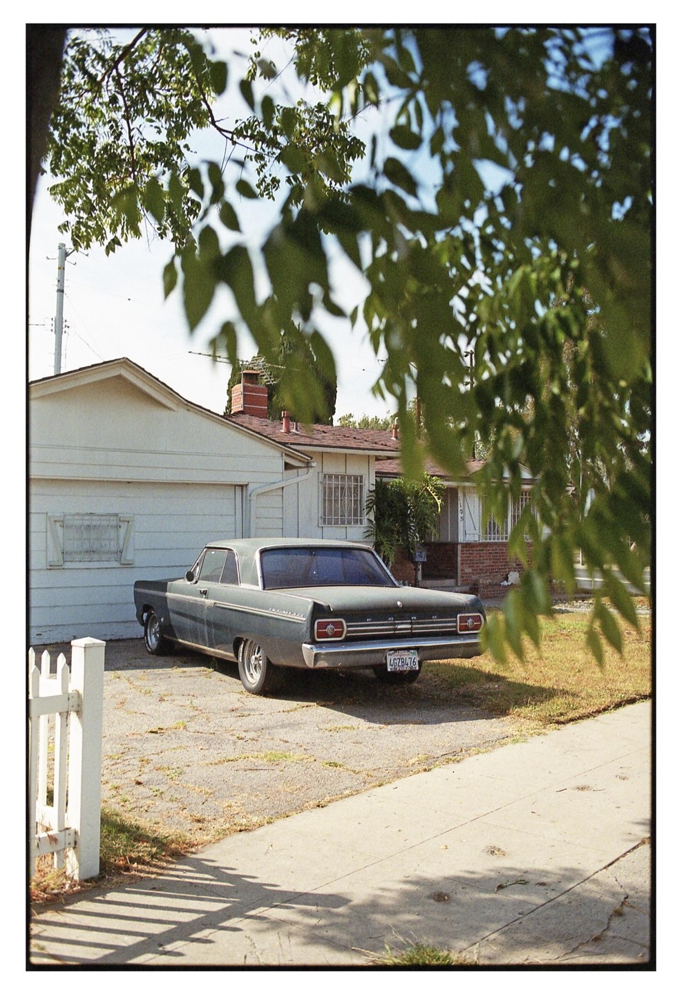 A vintage car is parked in a driveway next to a house, partially obscured by tree leaves in the foreground.