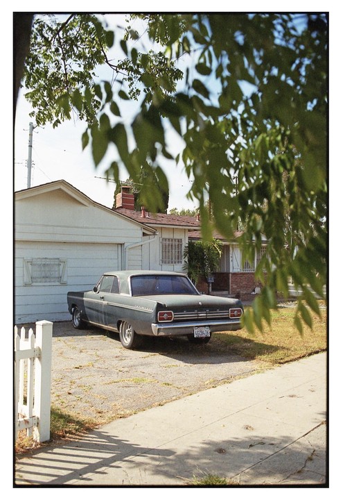 A vintage car is parked in a driveway next to a house, partially obscured by tree leaves in the foreground.