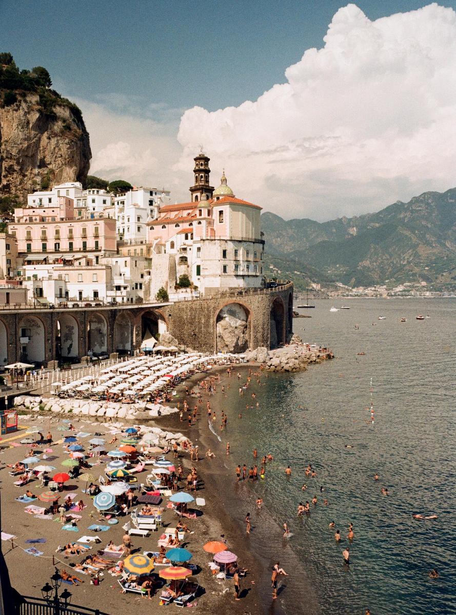 A coastal town with a church and many buildings built into a cliff overlooks a beach full of people and umbrellas.