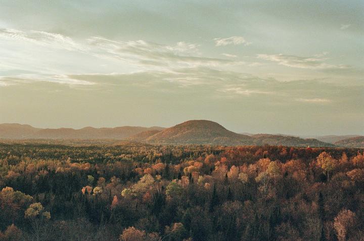 A wide landscape shows a forest with autumn foliage and hills under a cloudy sky.