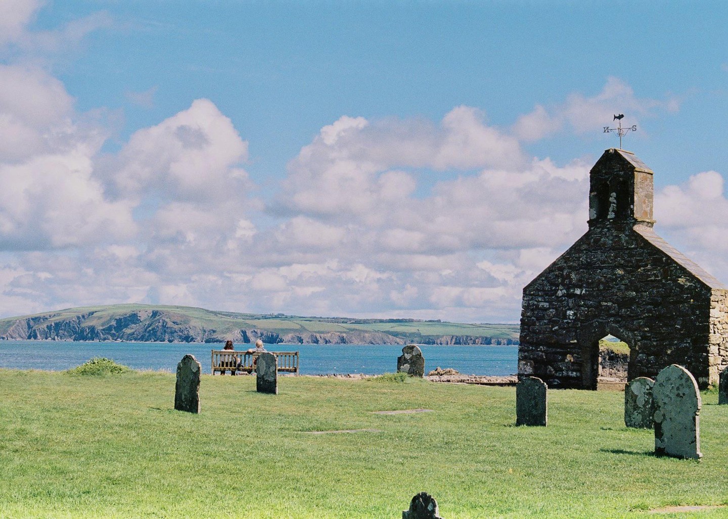 A stone church and gravestones stand on a grassy hill overlooking the ocean, with a couple sitting on a bench in the distance.