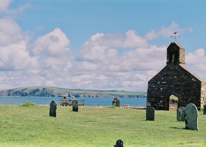 A stone church and gravestones stand on a grassy hill overlooking the ocean, with a couple sitting on a bench in the distance.