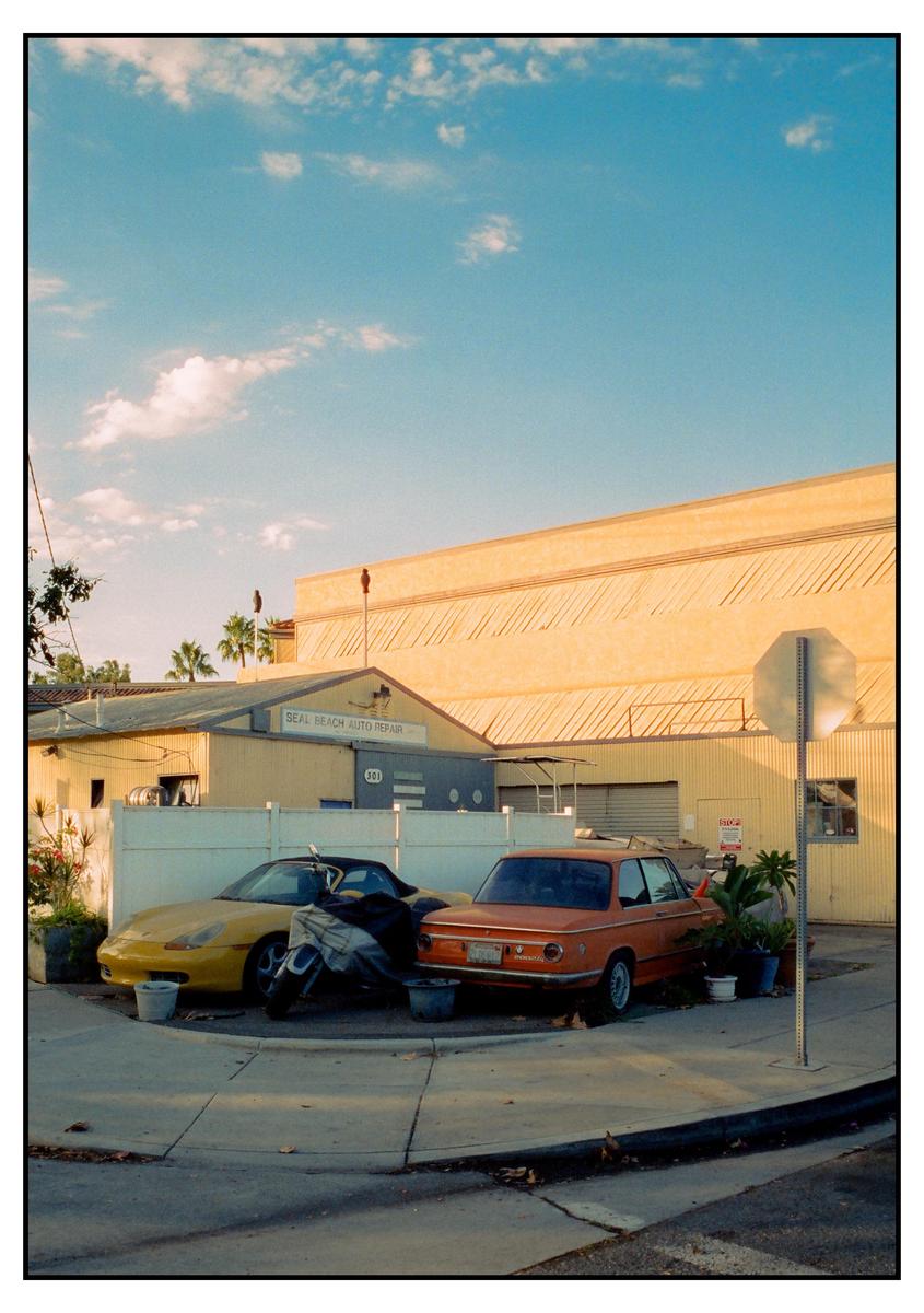 Two cars are parked in front of a body shop under a blue sky with clouds.