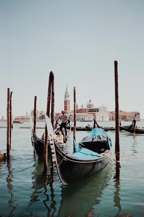 Several gondolas are moored to wooden posts in the water with a large building and bell tower in the background.