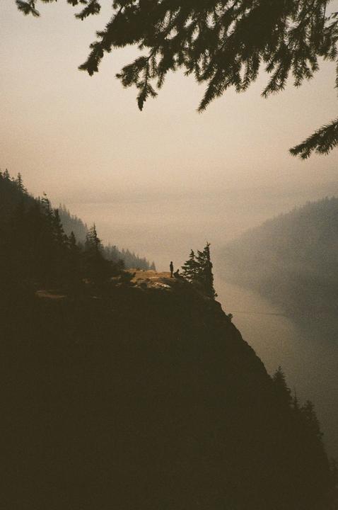 A person stands on a cliff overlooking a hazy valley with a river and forested mountains.