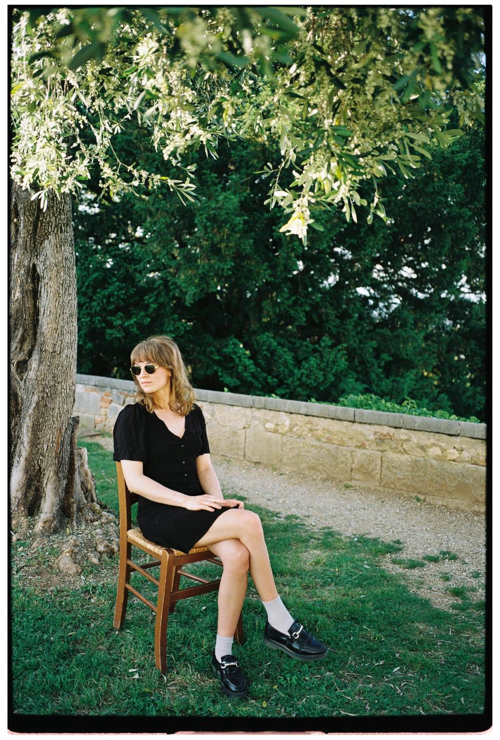 A woman in a black dress and sunglasses sits on a wooden chair under a tree.