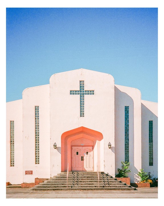 A pastel pink church with a cross-shaped window stands under a clear blue sky.