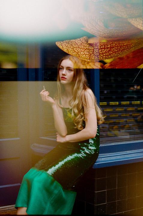 A woman in a green dress sits by a window, holding a cigarette, with reflections of leaves visible on the glass.