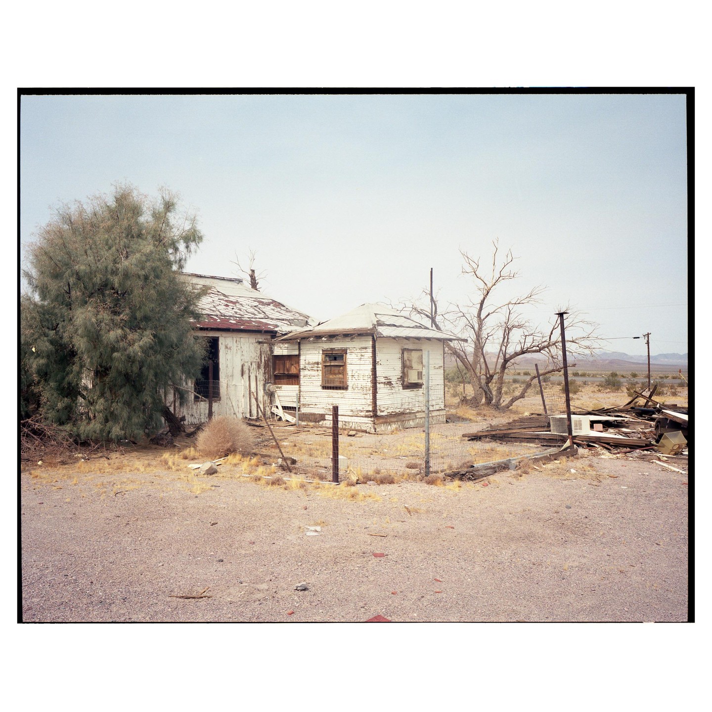 Two dilapidated buildings stand in a desert landscape with a large tree on the left and debris on the right.