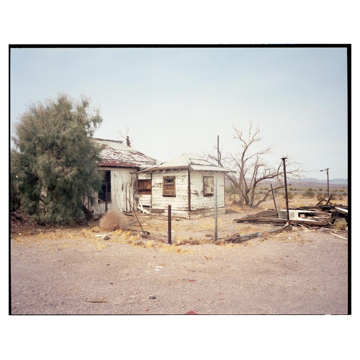 Two dilapidated buildings stand in a desert landscape with a large tree on the left and debris on the right.