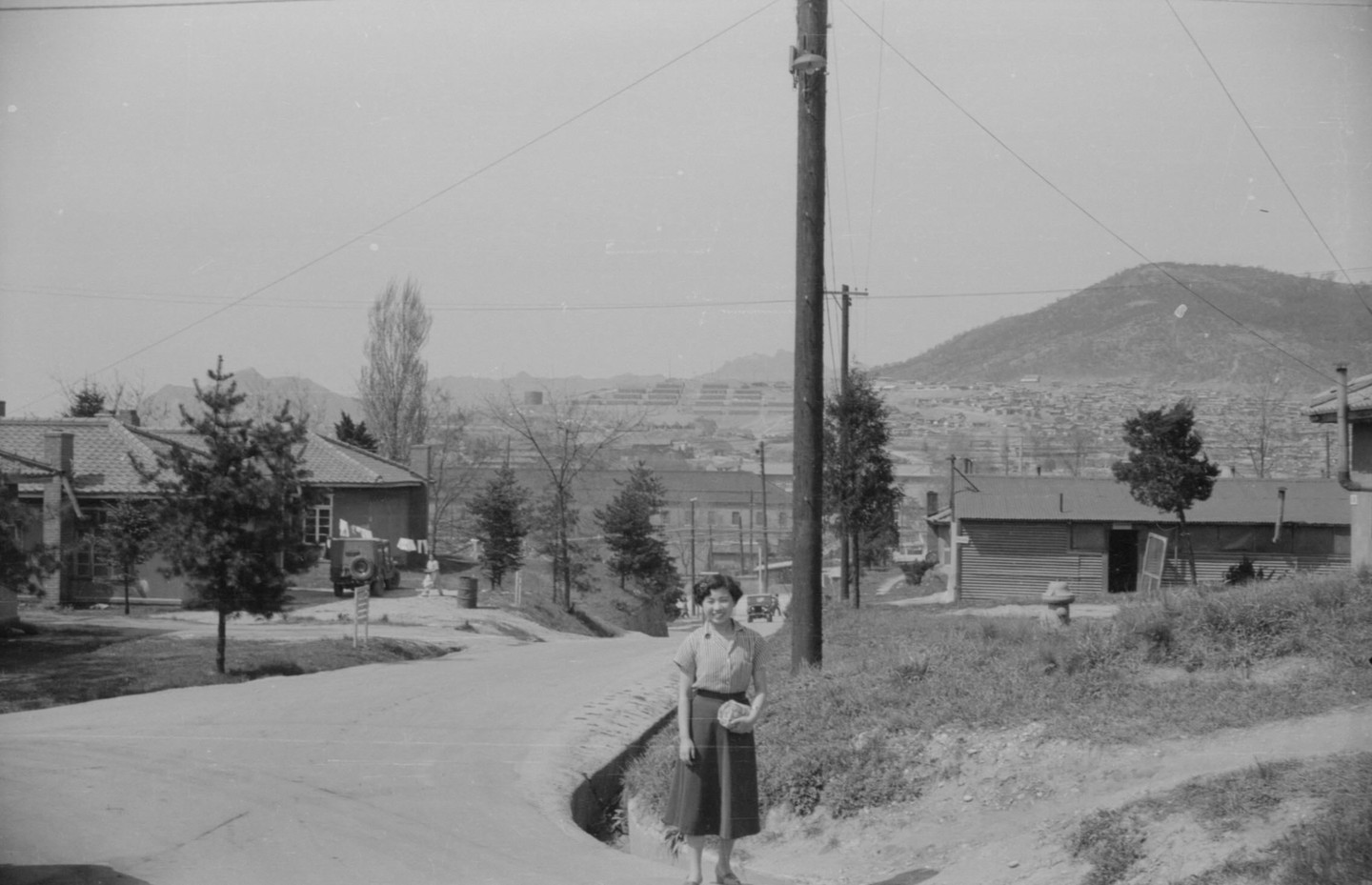 A woman stands on a dirt road with buildings and a mountain in the background.