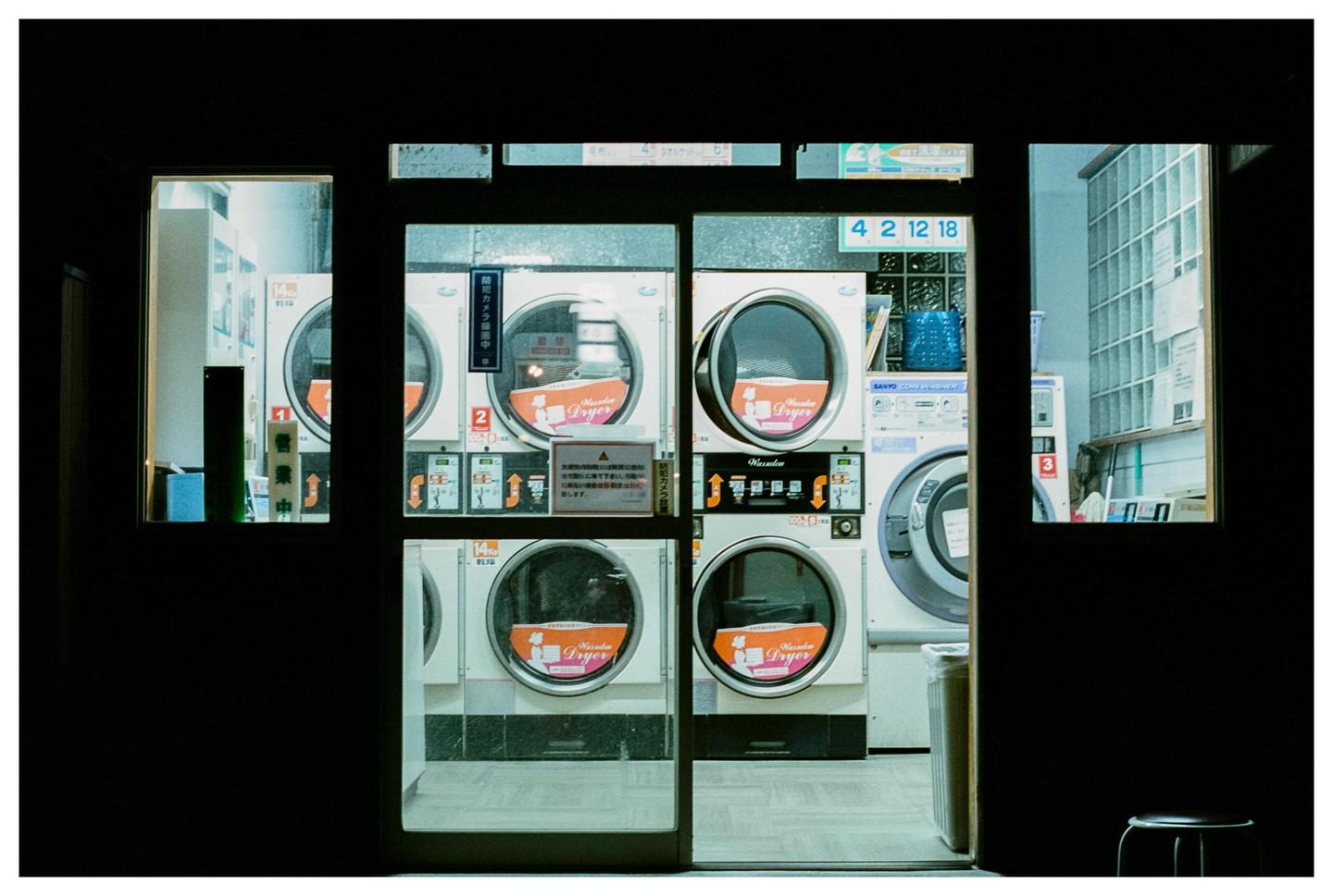 A row of washing machines and dryers are visible through the glass doors and windows of a laundromat at night.