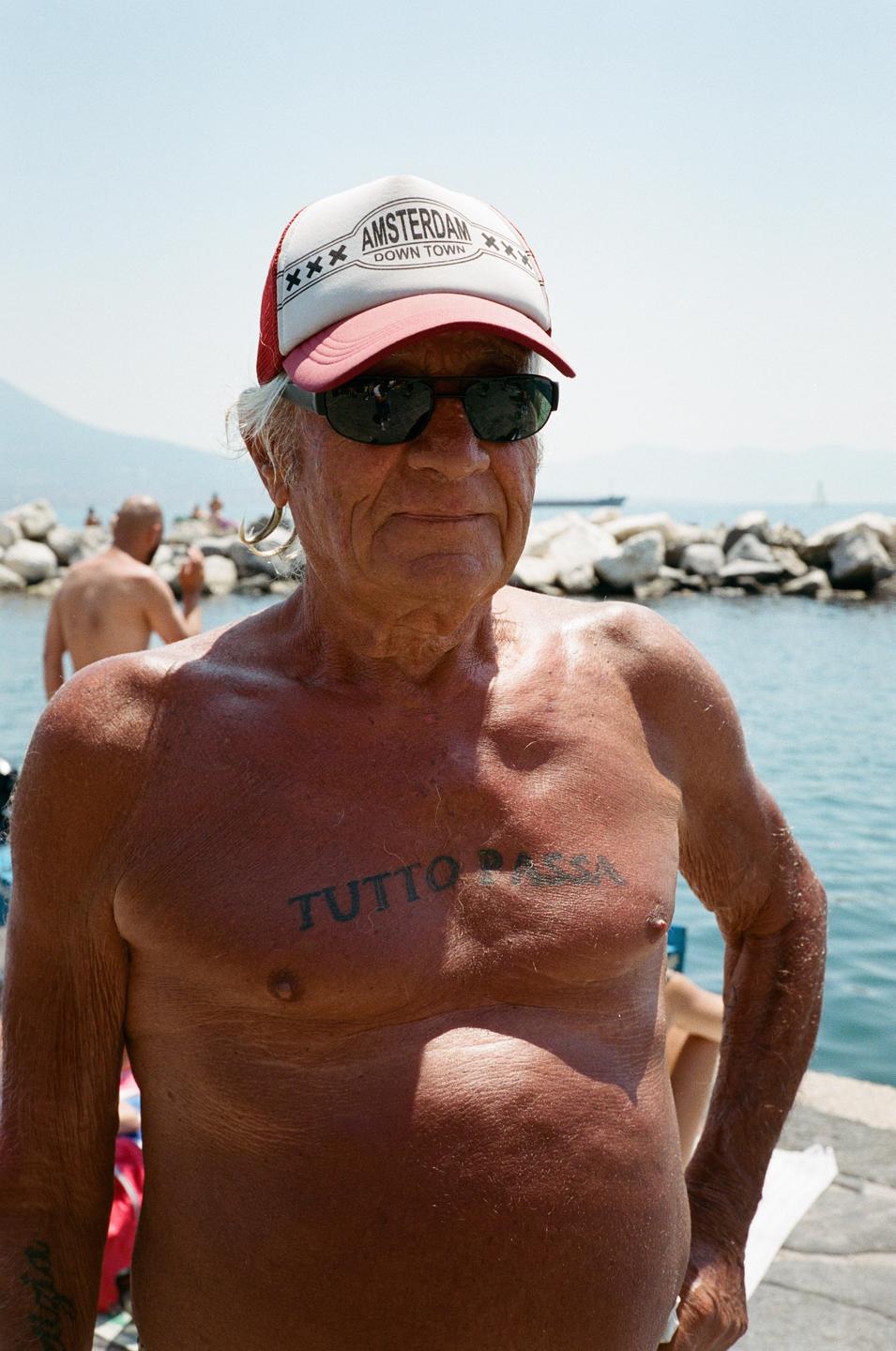 A man wearing a cap and sunglasses stands shirtless by the water with rocks and other people in the background.