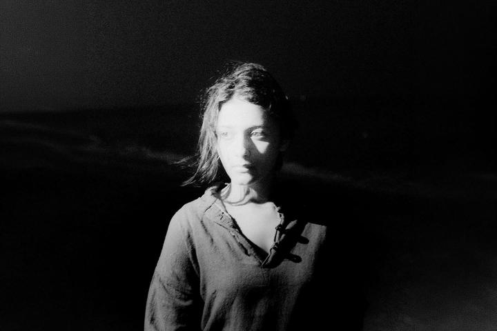 A woman with dark hair is illuminated by a strong light source from the left, casting a shadow across her face and body.