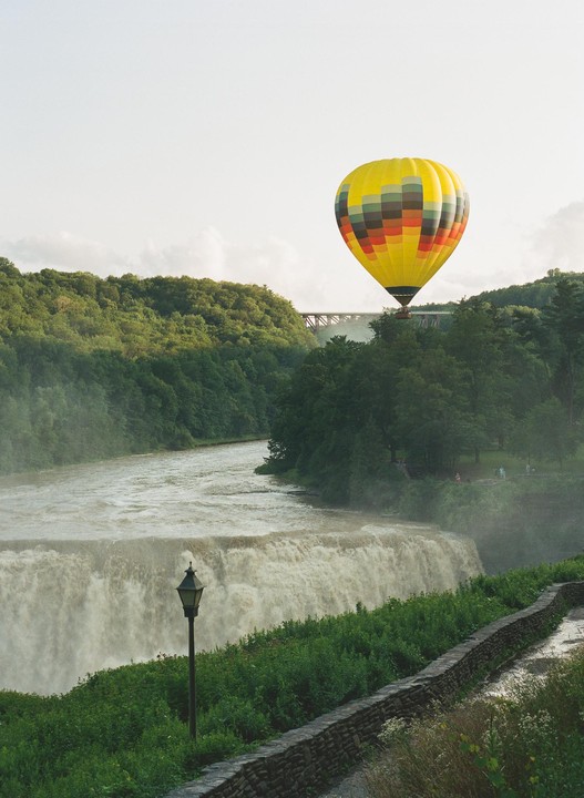 A yellow hot air balloon with colorful stripes floats above a waterfall and a forested gorge.