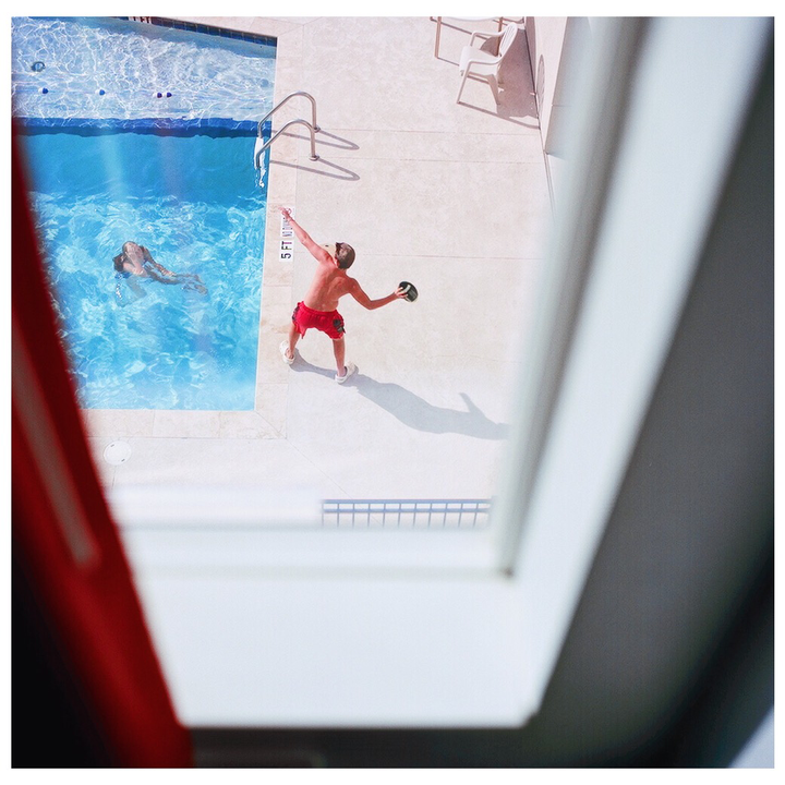 A boy in red shorts throws a football next to a swimming pool where a woman is swimming.