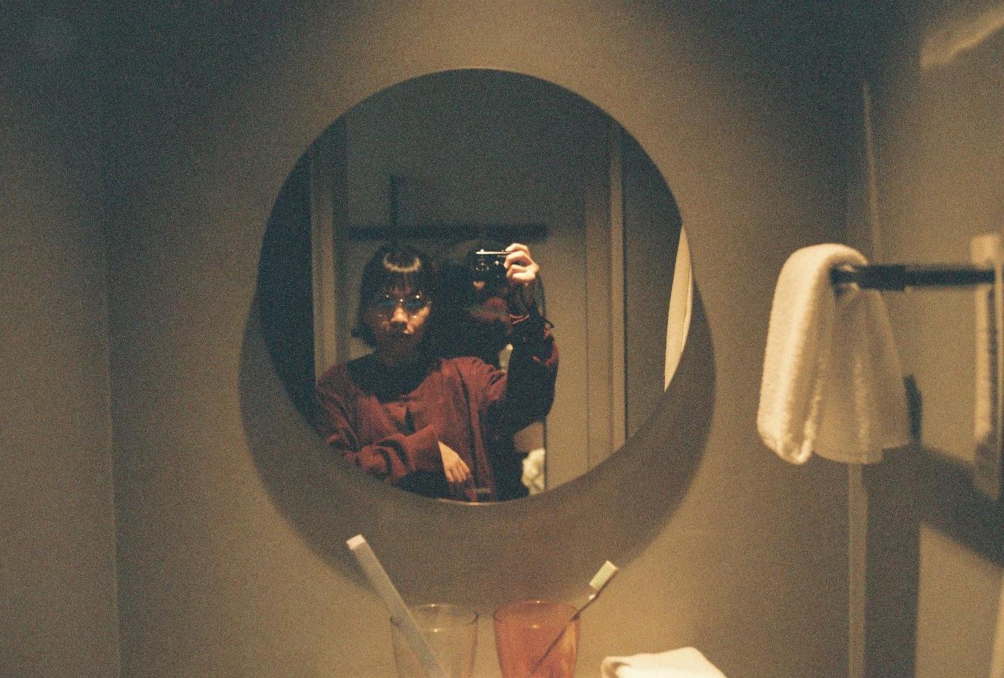 A woman with short hair and glasses takes a self-portrait in a round mirror in a hotel bathroom.