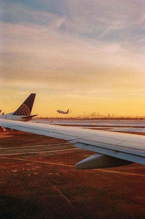 An airplane wing is visible in the foreground as another airplane takes off in the distance with a city skyline on the horizon at sunset.