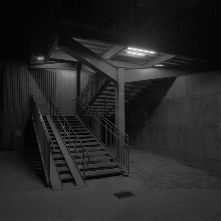 A dark, empty underpass features a set of stairs with a ramp and a second set of stairs leading upwards.