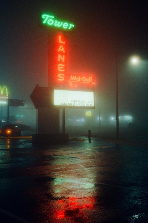 A neon sign for Tower Lanes and Mini-Golf glows through thick fog at night, reflecting on the wet pavement.