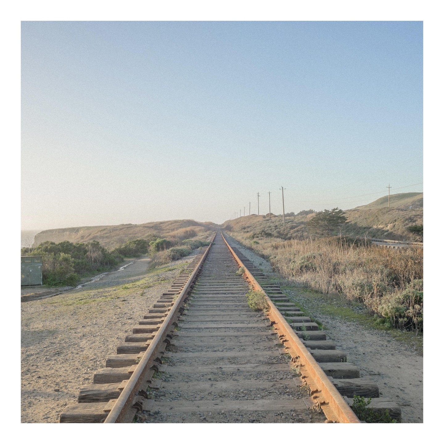 A long, straight railroad track extends into the distance, flanked by dry grass and hills under a clear sky.