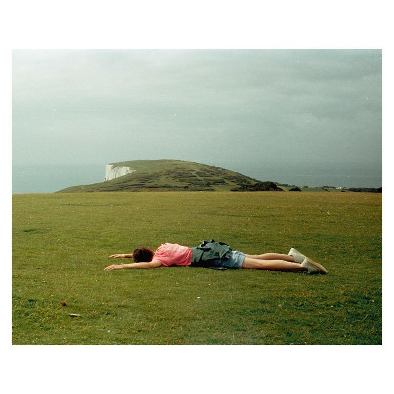 A woman lies face down on a grassy hill overlooking a body of water and a white cliff.