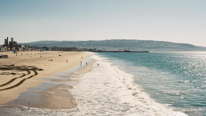 A wide view of Hermosa Beach shows waves breaking on the shore, with buildings and hills in the distance.