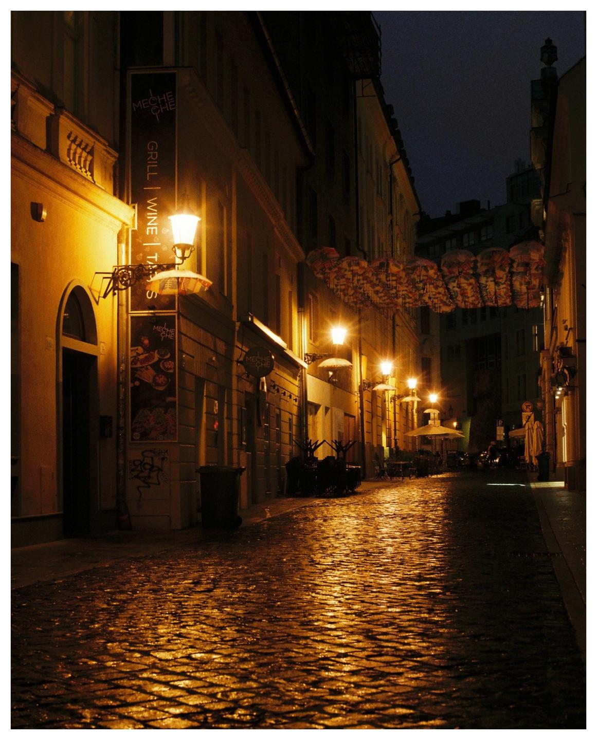 A narrow cobblestone street is illuminated by warm streetlights at night, with reflections visible on the wet ground.