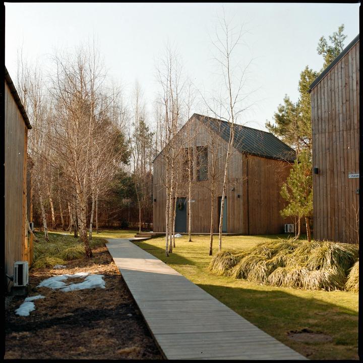 A wooden walkway leads to a modern wooden house surrounded by trees and patches of snow on the ground.
