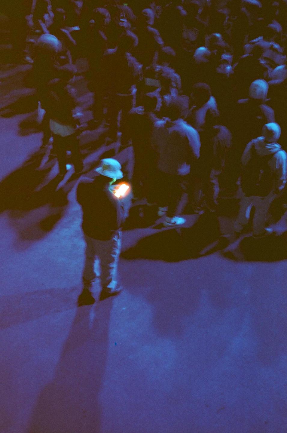 A person in a bucket hat holds a glowing object while standing in a crowd at night.
