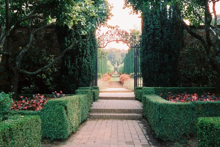 A brick path leads through a formal garden with hedges, trees, and a decorative iron gate.