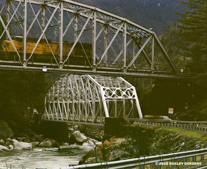 A yellow train crosses a metal bridge that spans over another metal bridge and a river.