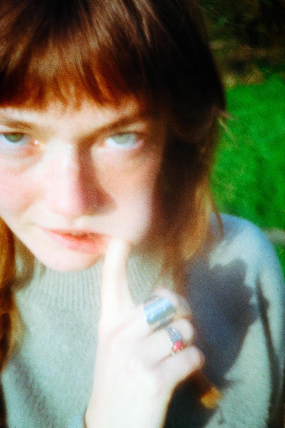 A woman with red hair and green eyes looks directly at the viewer, with her finger touching her lips.