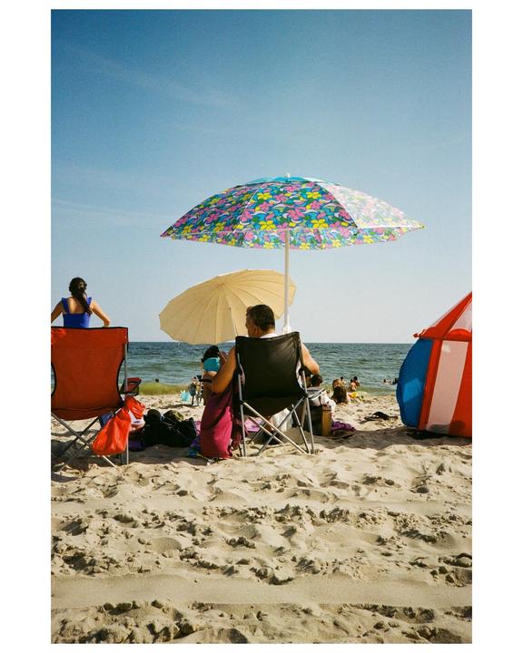 A man and a woman sit in beach chairs under colorful umbrellas on a sandy beach with the ocean in the background.