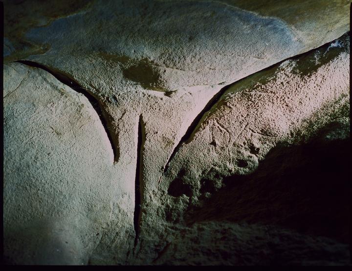 A close-up shot of a cave wall shows a natural rock formation resembling a vulva, with carvings on the right side.