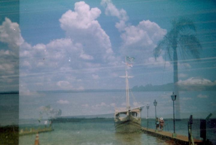 A boat with a flag is docked at a pier with two people and a palm tree reflection.