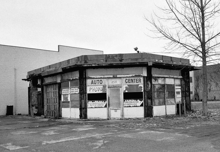 An abandoned auto center with broken windows and a bare tree stands under a cloudy sky.