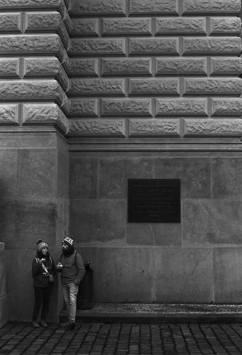 A couple stands on a cobblestone street next to a stone building with a plaque.