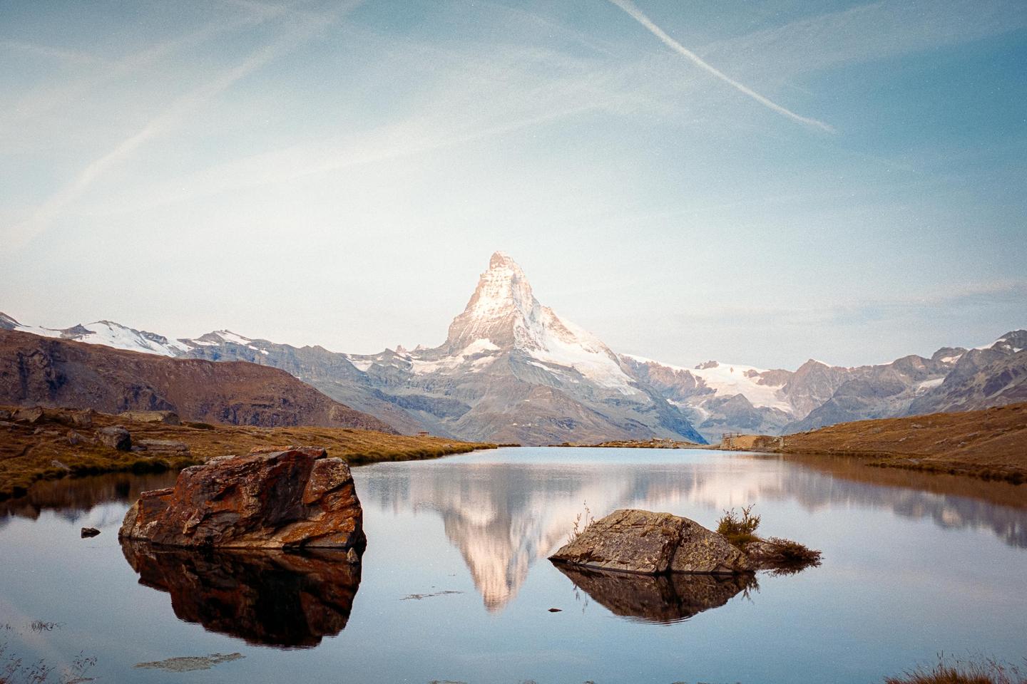 A snow-capped mountain peak is reflected in a still lake with rocky shores under a bright sky.