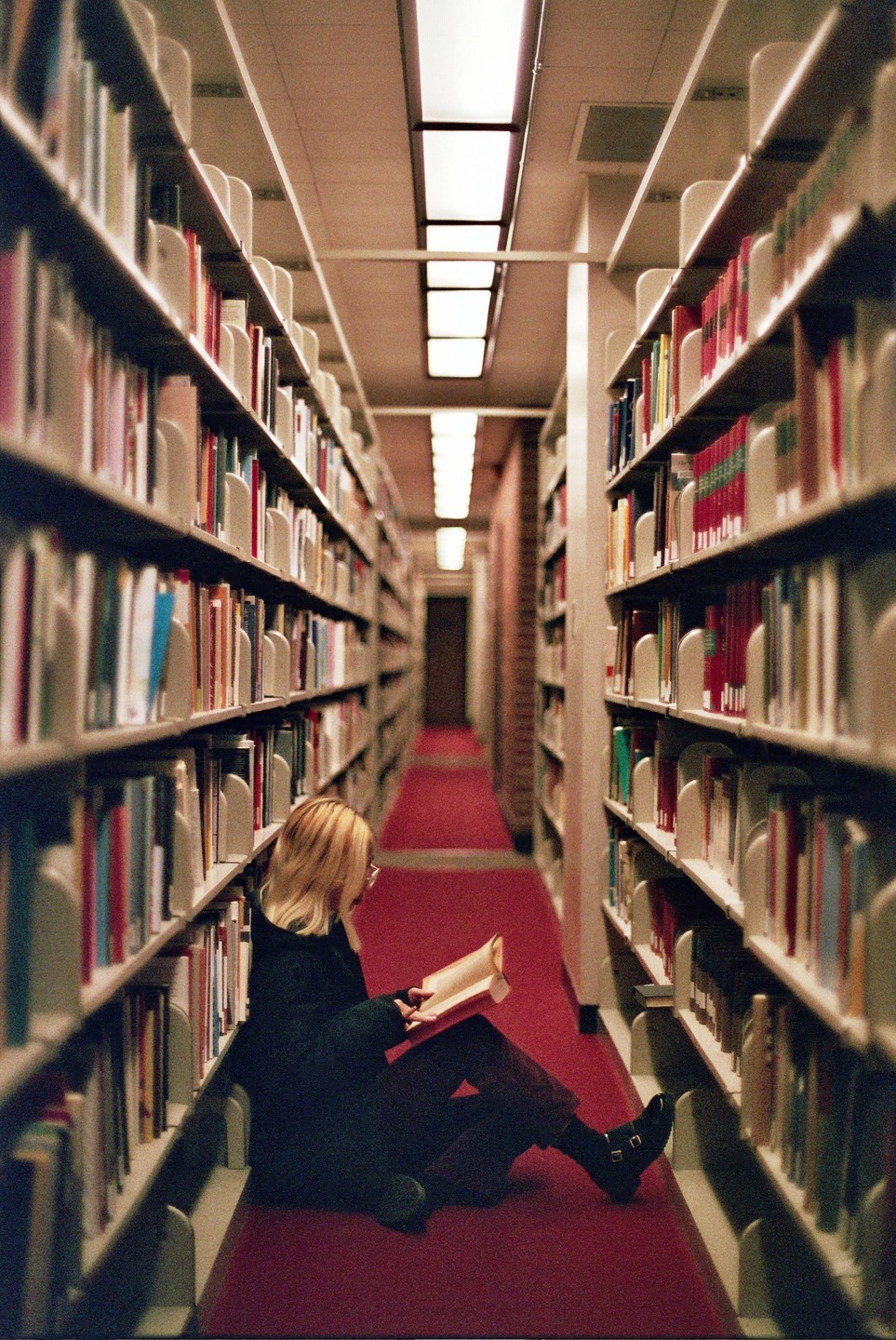 A woman sits on a red carpeted floor between two tall bookshelves and reads a book.