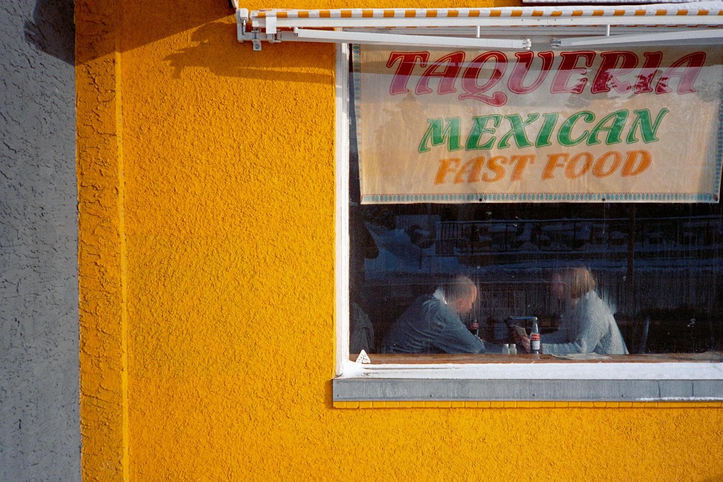 Two people sit inside a taqueria with a sign for Mexican fast food visible in the window.