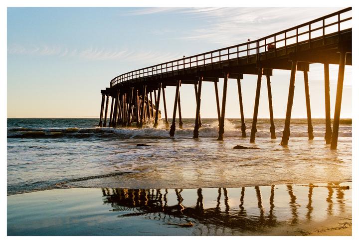 A long pier curves over the ocean with waves crashing against its supports at sunset.