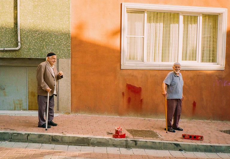 Two elderly men stand on a brick sidewalk next to a building with an orange wall and a green tiled wall.
