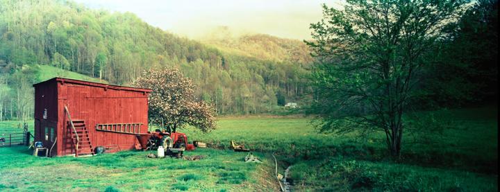 A red barn stands in a green field with a blooming tree and a tractor, with a forest and mountains in the background.