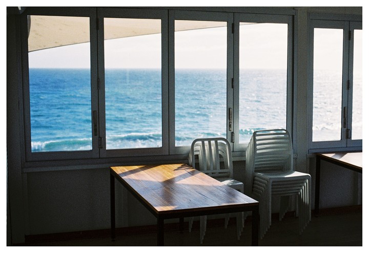 A wooden table and stacked chairs sit in a room with large windows overlooking the ocean.