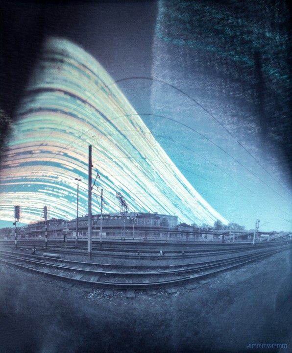 A long exposure photograph shows a train station with multiple tracks and buildings under a sky with light trails.