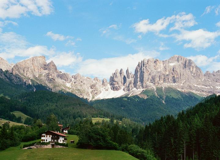 A house with a red roof sits on a green hill in front of a forest and a range of rocky mountains.