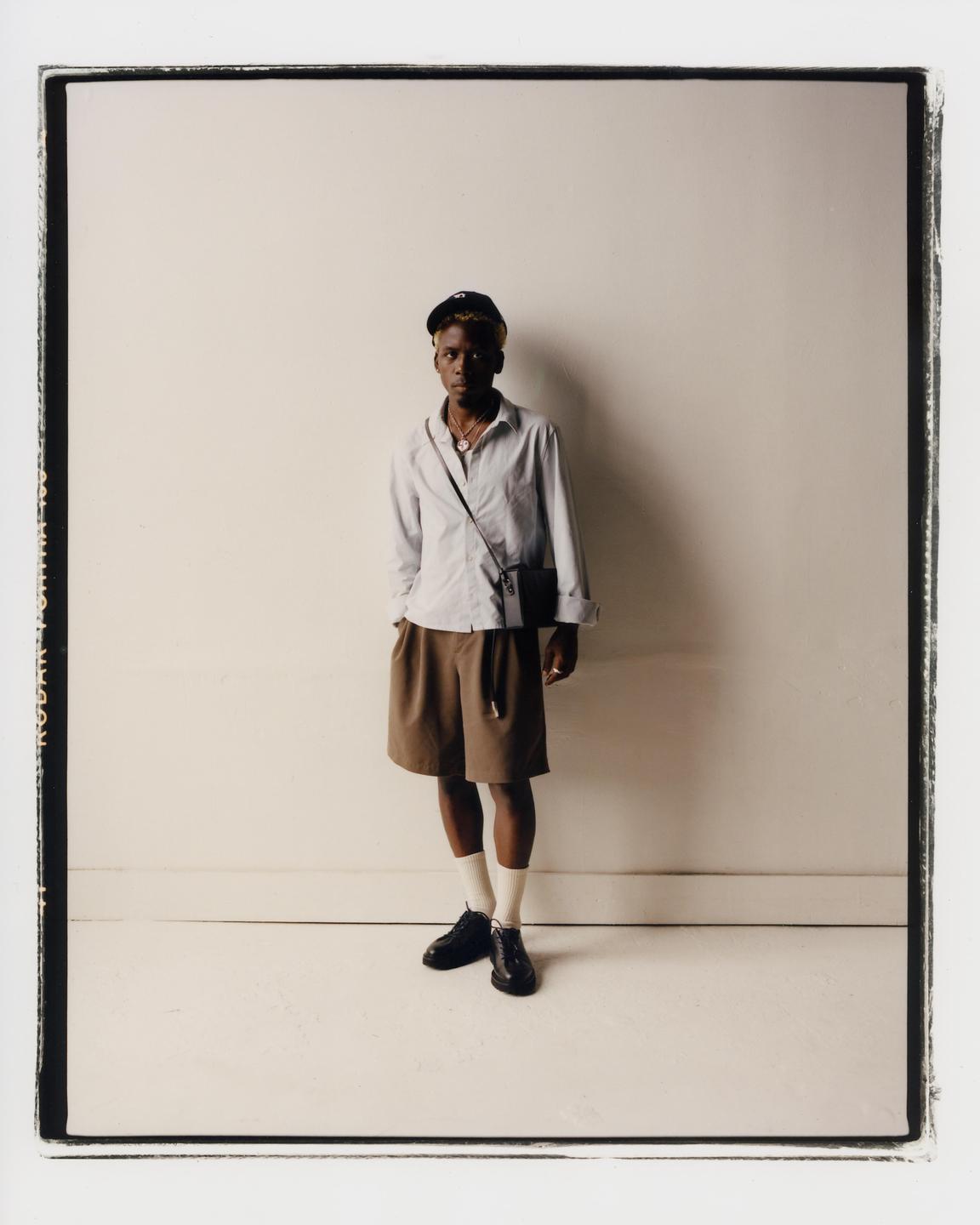 A man wearing a cap, shirt, shorts, and shoes stands against a white wall.